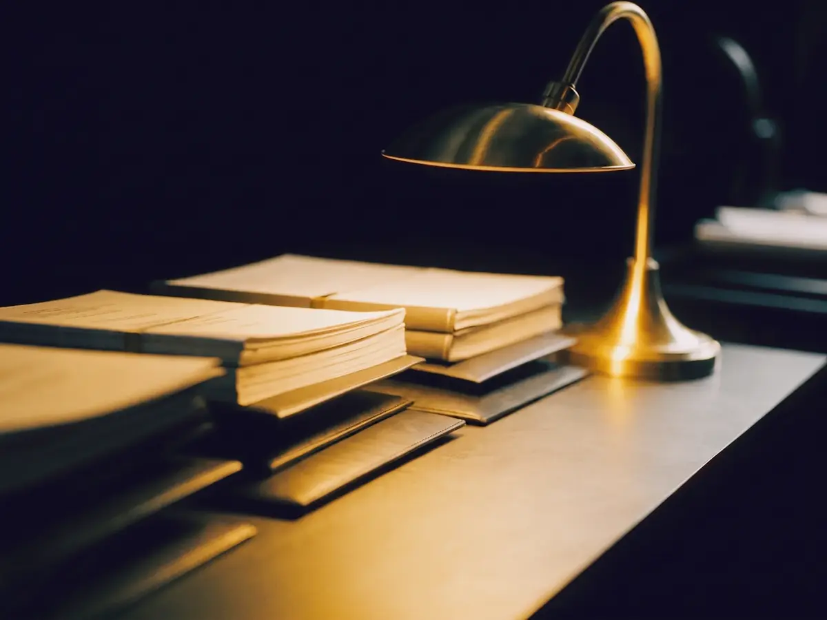 Business filing documents and closed folders stacked on a dark desk beside a lit brass lamp